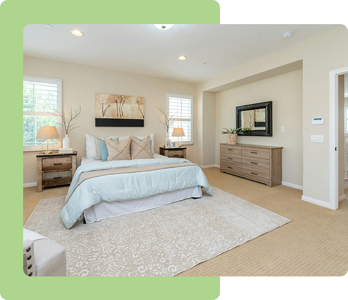 A spacious bedroom with a sustainable mattress, two bedside tables, a dresser with a mirror, lamps, and wall art. The room has beige walls, a large rug, and windows with white shutters.